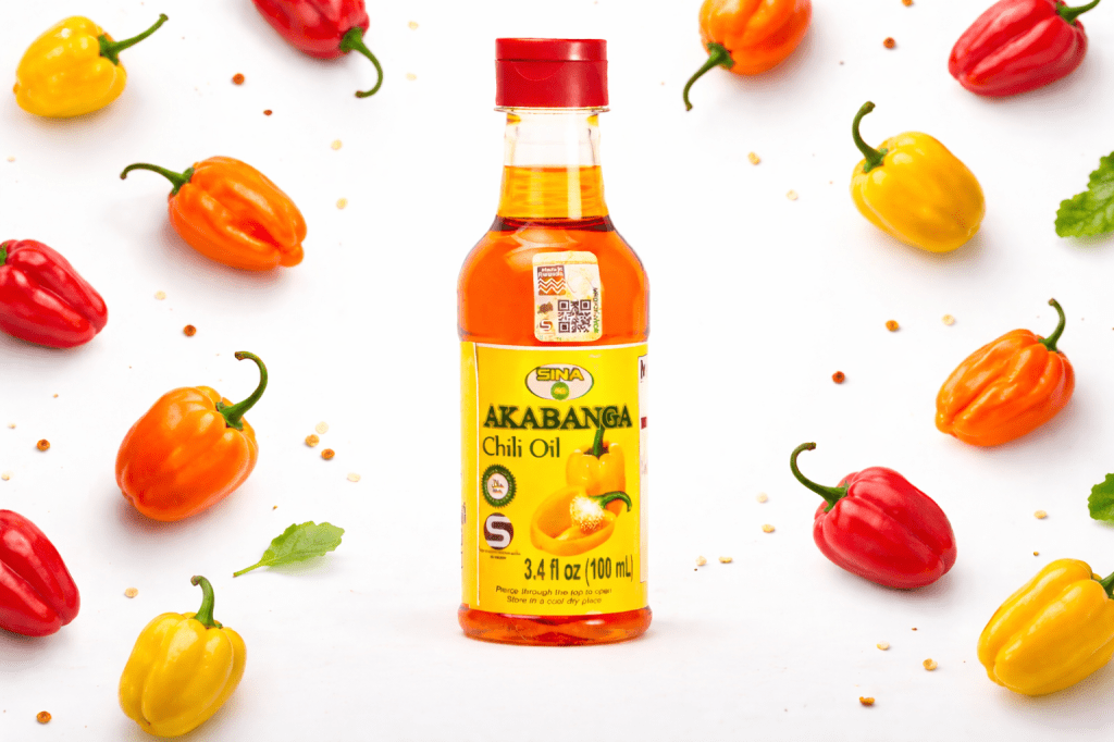 Bouteille d'huile de chili 'Akabanga' entourée de piments rouges, jaunes et oranges sur fond blanc.